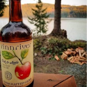 Finn River Oak and Apple Cider On a Picnic Table with a Woodland Scene Behind 