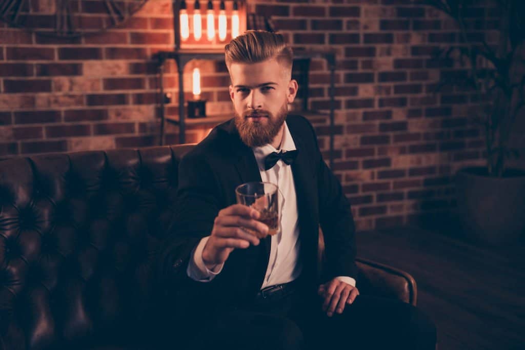 Joking hipster sitting on a leather couch cheersing with a whiskey