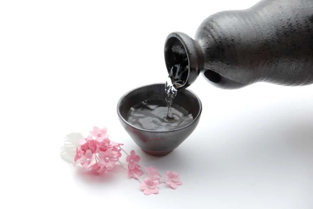 Sake pouring from a black ceramic carafe into a black ceramic cup with cherry blossoms on a white table
