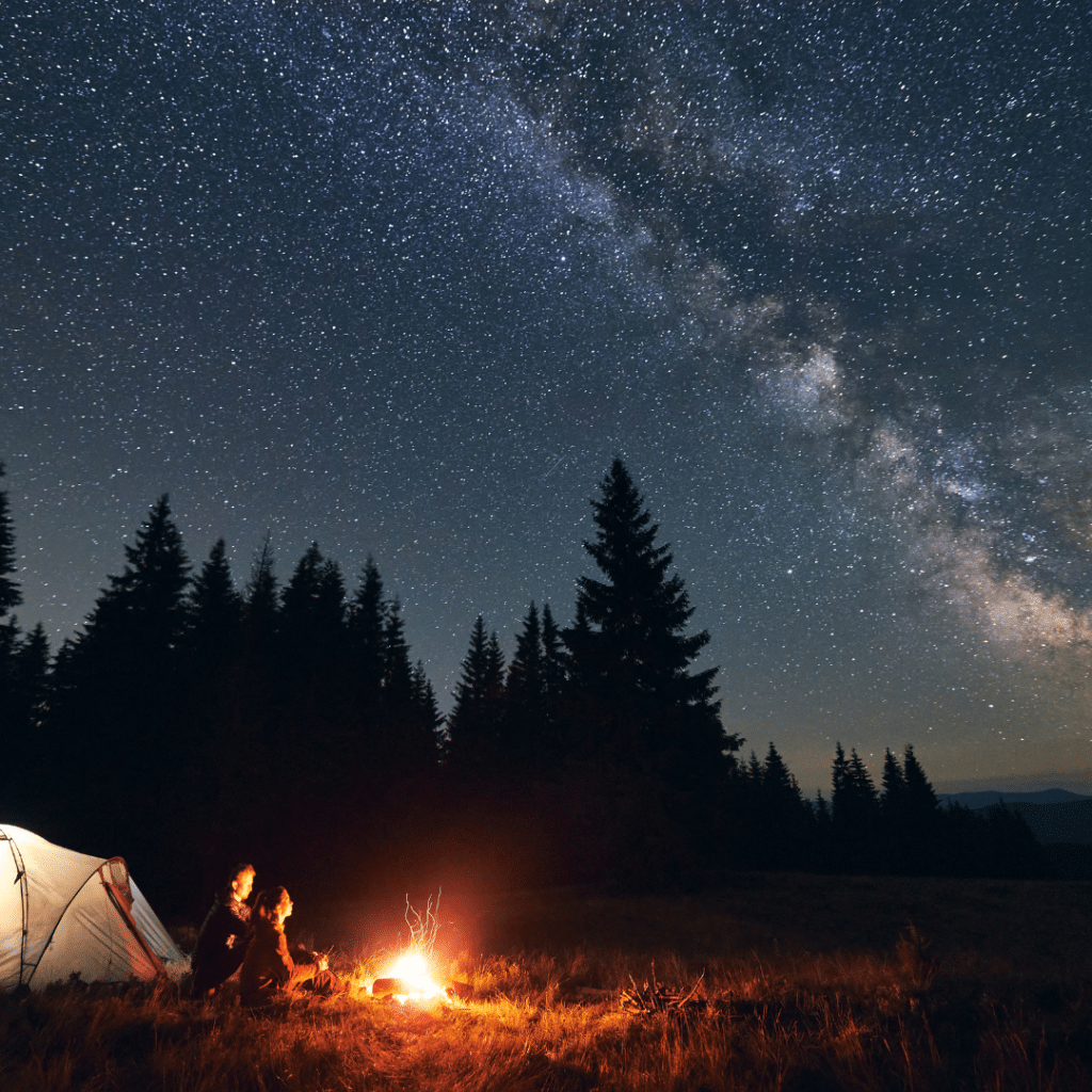 Image of a couple sitting around a campfire with the stars above. Great for camp cocktails!